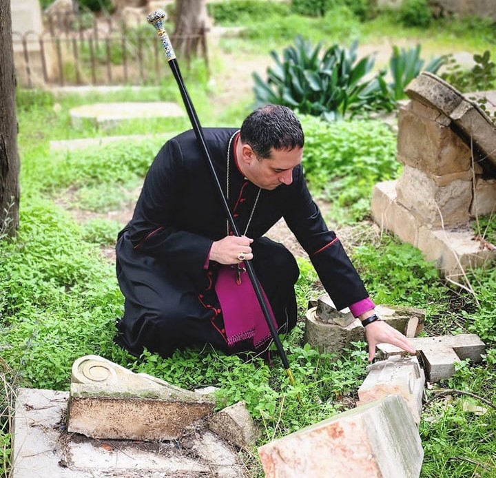 Archbishop Hosam bending down to inspect a broken cross from a grave