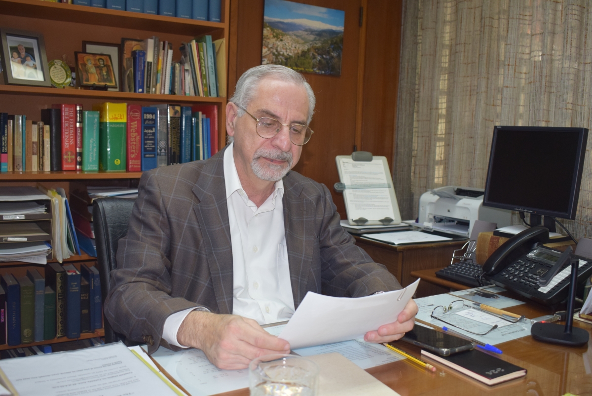 Dr George Sabra sat reading a paper at his desk in front of a large, full bookshelf