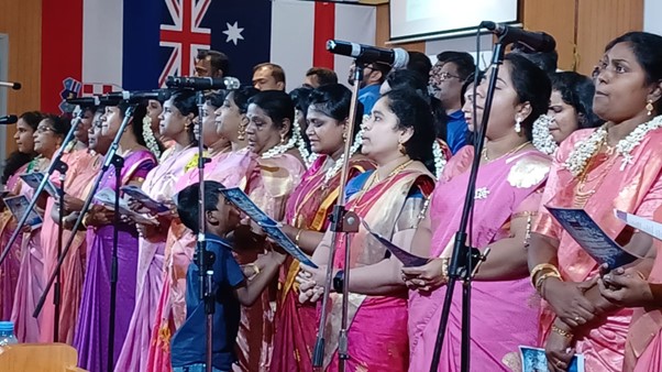 The choir behind the microphones the women wearing pink and purple dresses and the men in blue