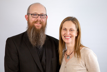 Fr Kevin with a long beard and wearing a black suit and glasses along with his wife smiling by his side with long light brown hair