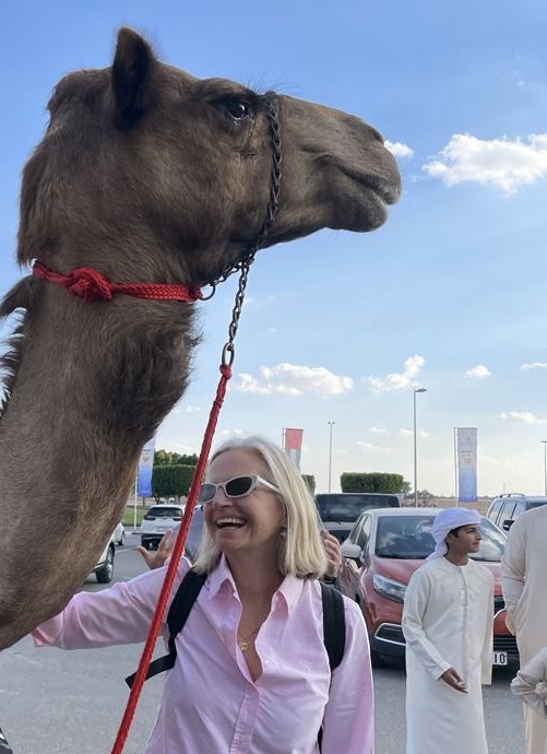 Christine, dressed in a pink blouse, sunglasses with a rucksack on her back, stroking to a camel's neck