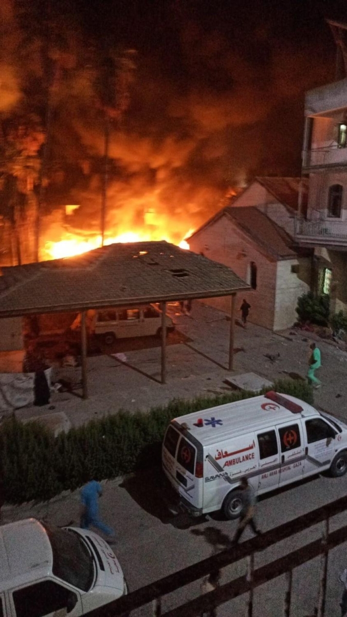View of the burning hospital from a balcony from across the road, with ambulances and people running